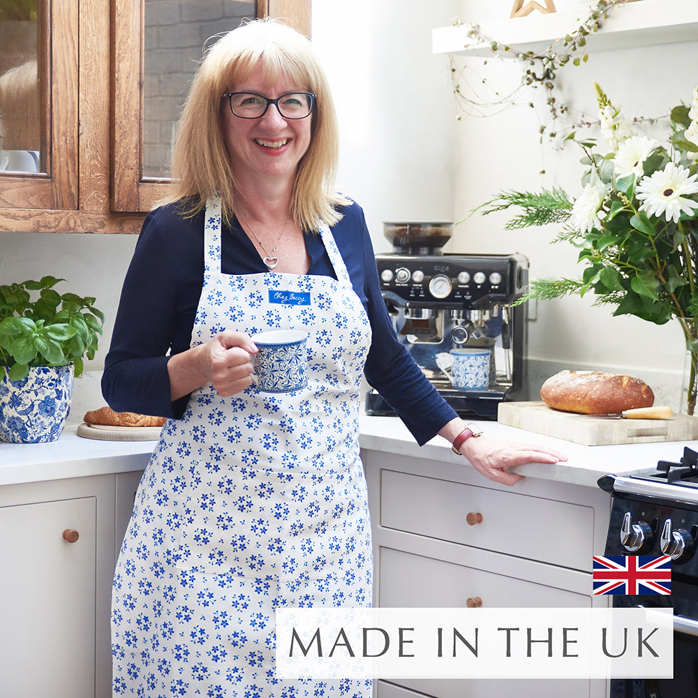 Beccy wearing the Blue Ditsy Floral Apron in a kitchen, holding a cup of tea, with a hand-drawn forget-me-not floral design on 100% half panama cotton