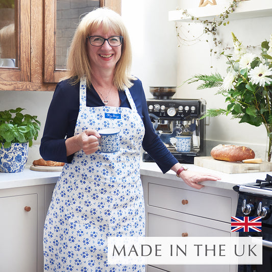 Beccy wearing the Blue Ditsy Floral Apron in a kitchen, holding a cup of tea, with a hand-drawn forget-me-not floral design on 100% half panama cotton