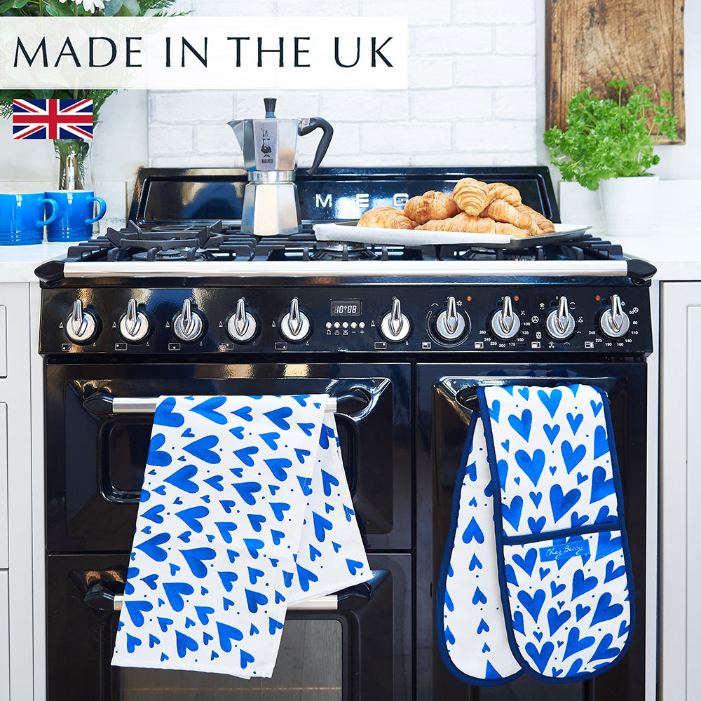 Blue hearts tea towel hanging on a black range cooker, accompanied by a matching double oven glove; a coffee pot rests atop the cooker beside a tray of freshly baked croissants.