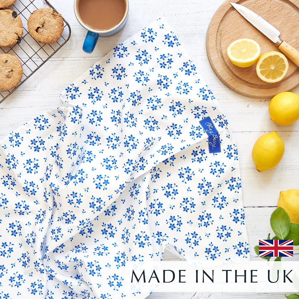 Chez Beccy Blue Ditsy Floral Tea Towel on a white wooden background, with a cup of tea, homemade biscuits on a wire rack, fresh lemons, and a small round wooden chopping board with a cut lemon.