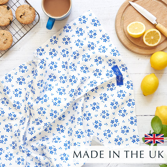 Chez Beccy Blue Ditsy Floral Tea Towel on a white wooden background, with a cup of tea, homemade biscuits on a wire rack, fresh lemons, and a small round wooden chopping board with a cut lemon.