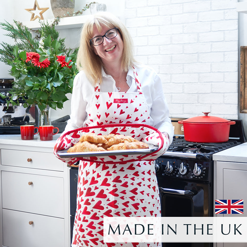 Beccy wearing the Red Shooting Heart Apron, holding a tray of freshly baked croissants in a kitchen. The apron features a bold red and white shooting heart design.