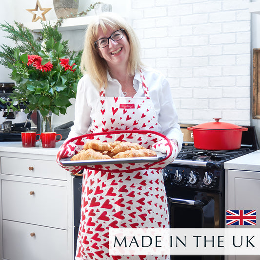 Beccy wearing the Red Shooting Heart Apron, holding a tray of freshly baked croissants in a kitchen. The apron features a bold red and white shooting heart design.