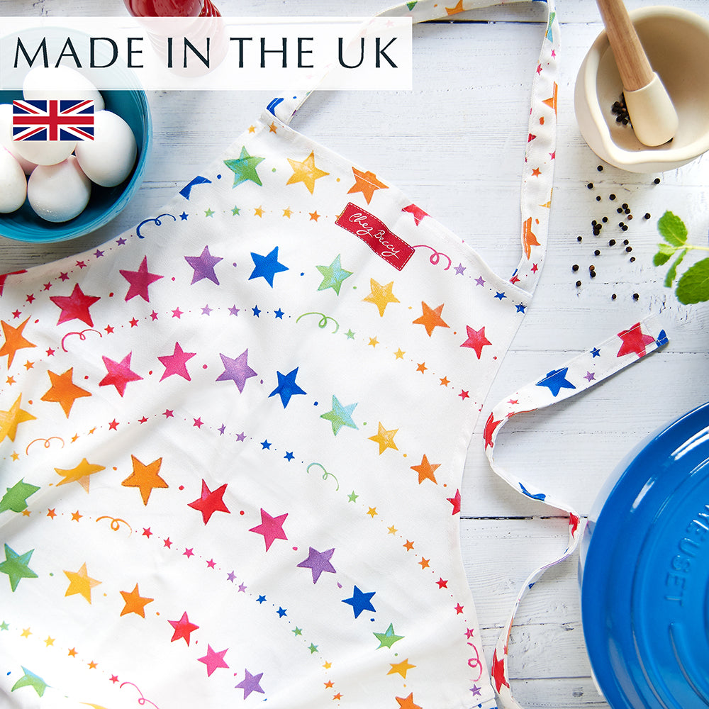 Rainbow Stars Apron flat lay on white wooden background, featuring a blue Le Creuset pot, pestle and mortar, and a bowl of eggs, with vibrant multi-coloured star design and extra-long fabric ties