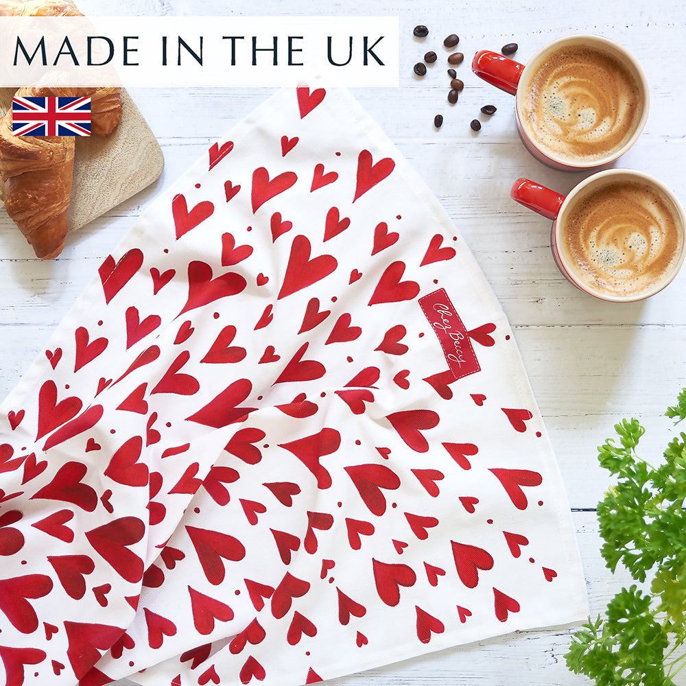 Red Shooting Hearts Tea Towel featuring a hand-painted design of red hearts on 100% half panama cotton fabric on a white wooden background with 2 cups of coffee and croissants. Made in the U.K.