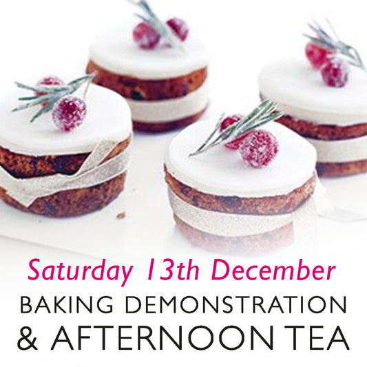 A promotional image for a baking demonstration and afternoon tea event on Saturday, 13th December. The image features beautifully decorated mini Christmas cakes, topped with white icing, sugared cranberries, and rosemary sprigs, wrapped with a festive ribbon. The text is styled in a mix of elegant black and festive pink fonts.