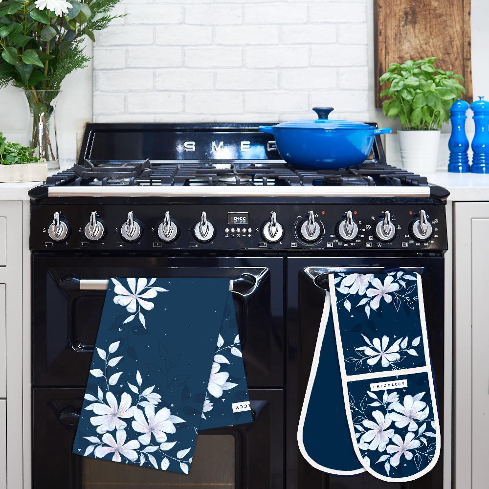 A stylish kitchen scene featuring a Navy Magnolia Floral Tea Towel and matching oven gloves displayed on a black range cooker. The luxury 100% half panama cotton tea towel and oven gloves showcase an elegant Magnolia-inspired floral design in soft white and blush tones on a deep navy background. A blue cast iron pot sits on the stove, complementing the timeless kitchen aesthetic. Perfect for adding elegance and practicality to any home.