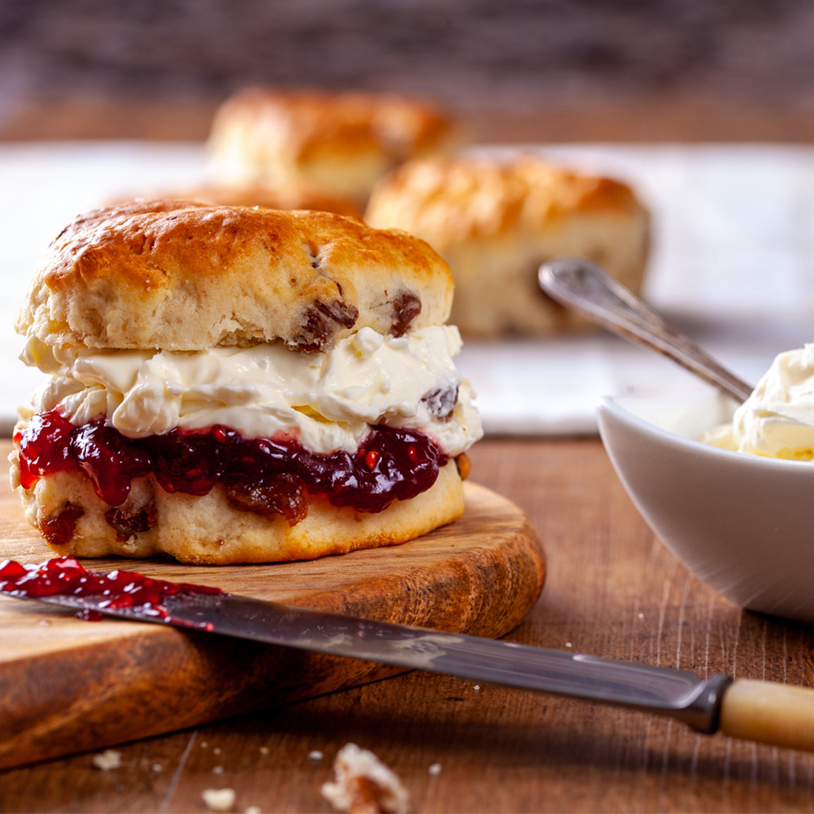 A freshly baked fruit scone, generously filled with clotted cream and raspberry jam, sits on a rustic wooden board. In the background, additional scones rest on a white cloth, and a small bowl of clotted cream with a spoon is nearby. A butter knife with jam rests on the board, adding to the inviting afternoon tea scene.