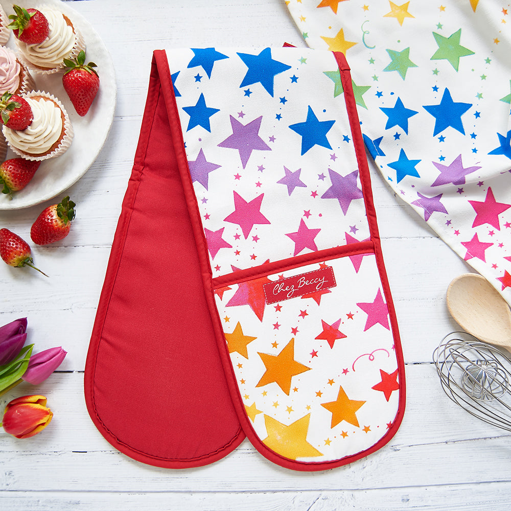 Rainbow shooting stars double oven glove with red trim, colourful cotton kitchen accessory by Chez Beccy on white wooden background with strawberries and tulips.