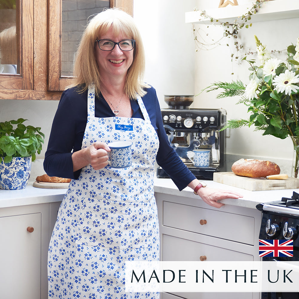 Beccy standing in a kitchen holding a cup of tea wearing a blue ditsy floral apron