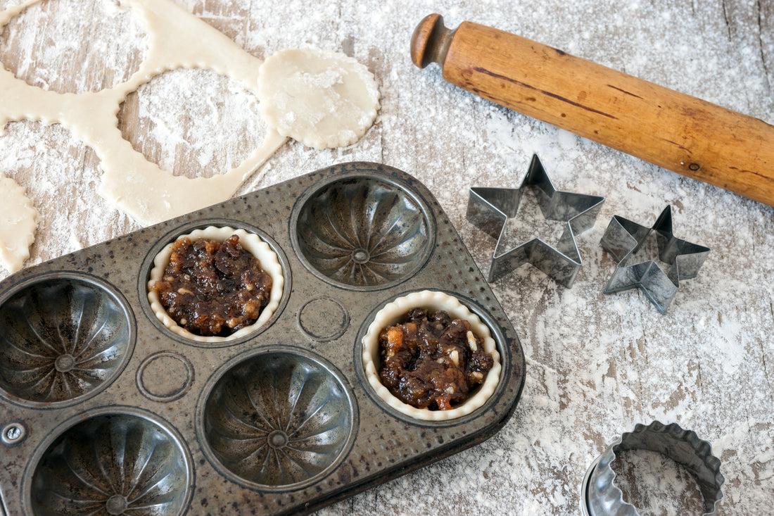 Unbaked mince pies in a baking tin with pastry cutters and rolling pin on a floured surface.