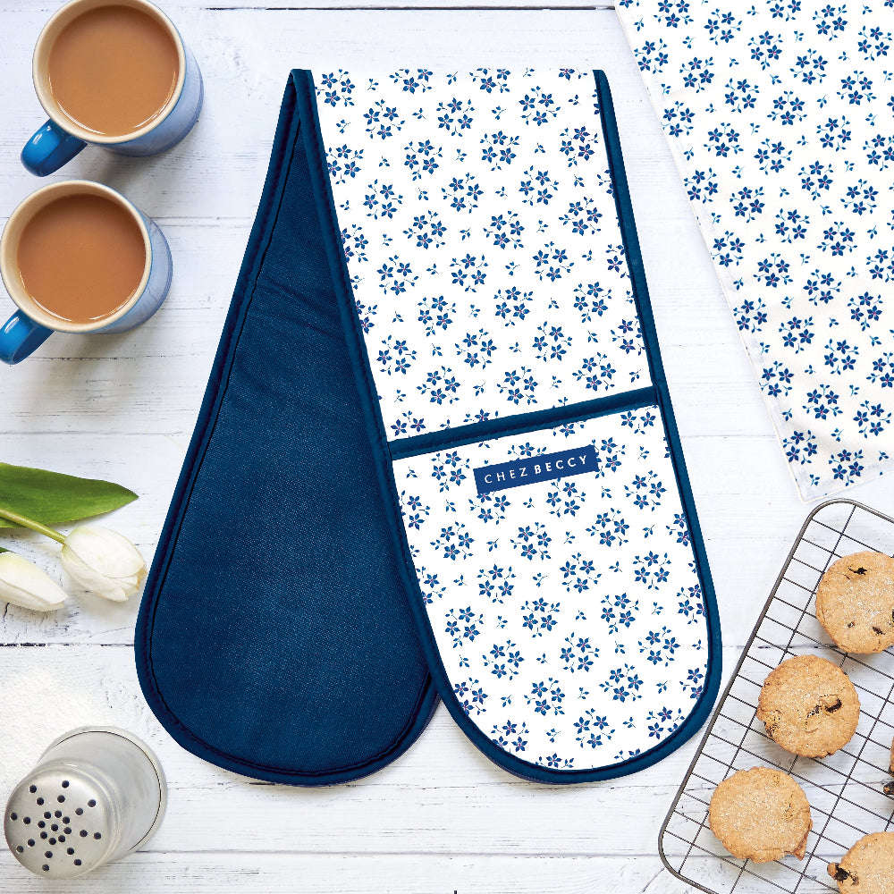 Chez Beccy Blue Ditsy Floral Double Oven Glove in a flatlay with cups of tea, tulips, cooling rack of biscuits, and matching kitchen textiles on a white wooden table.