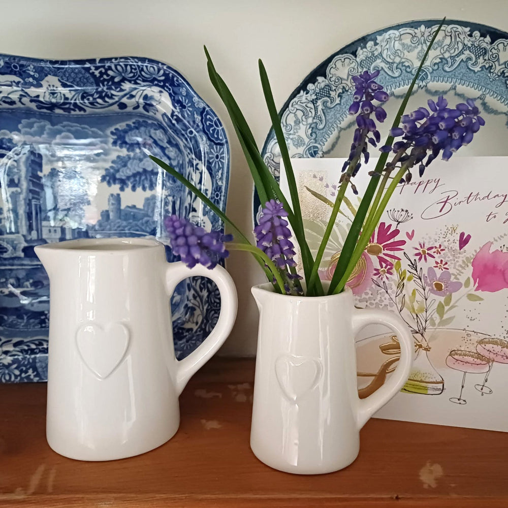 Two white ceramic pitchers with heart designs, one containing purple flowers, on a wooden surface with decorative plates and a birthday card in the background.
