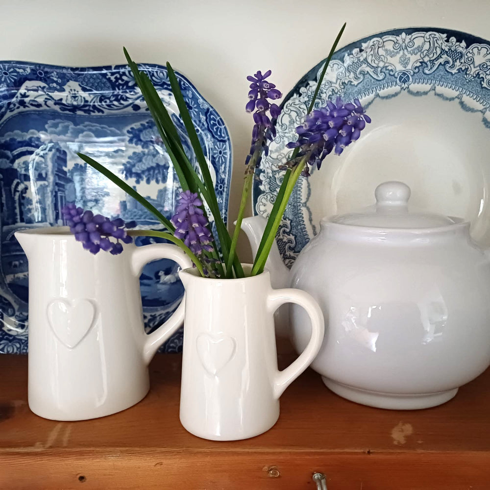 White ceramic pitcher with purple flowers on a wooden surface with blue and white patterned plates in the background.