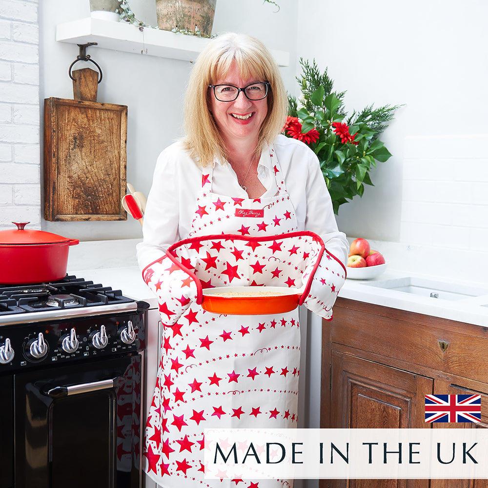 Beccy wearing the Red Shooting Stars Apron in the kitchen, holding a hot dish straight out of the oven, with a hand-drawn shooting star design in red and white