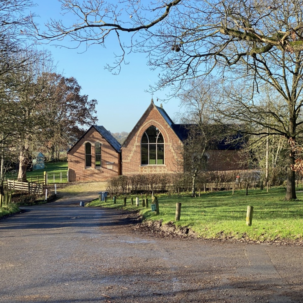 South Leigh village hall with a large arched window, situated in a grassy outdoor setting with trees and a paved road in the foreground.