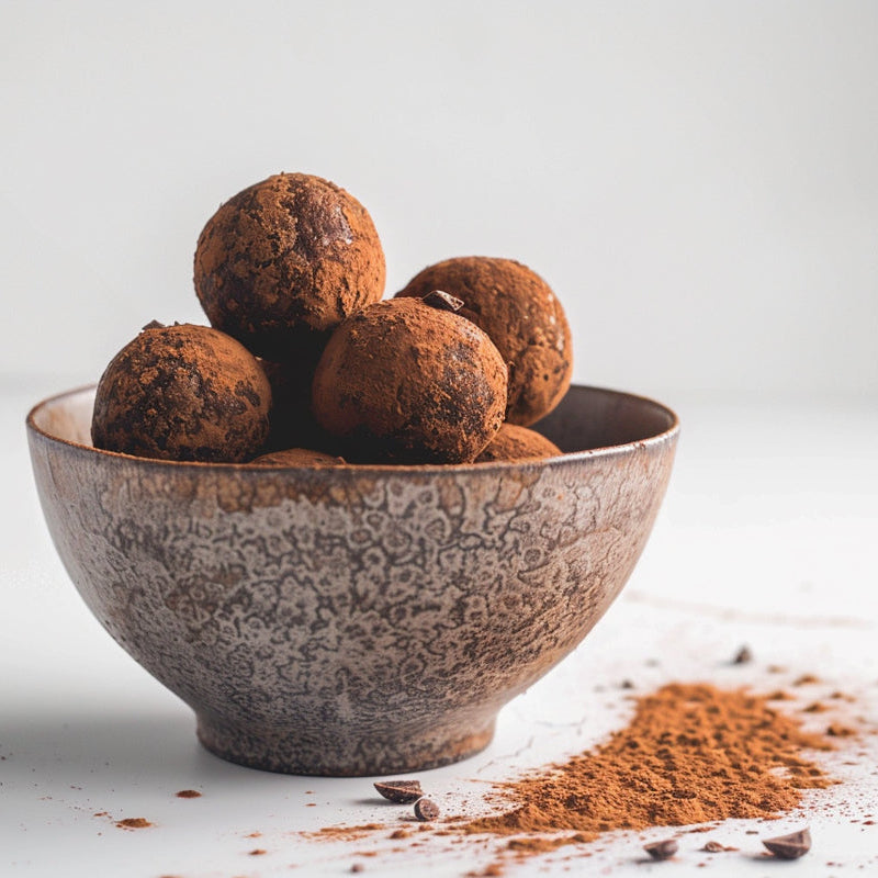 Five chocolate truffles dusted with cocoa powder are stacked in a textured ceramic bowl with a small pile of cocoa powder scattered on a light gray surface.
