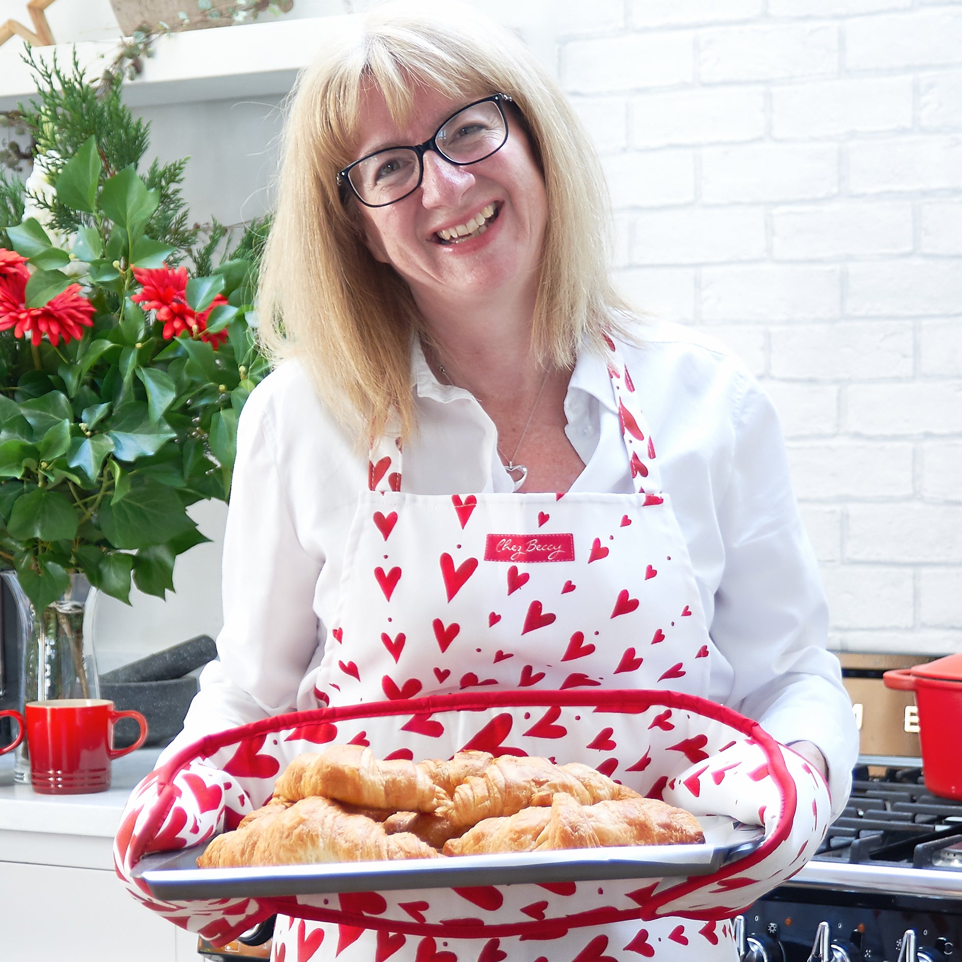 Cook Beccy in her kitchen holding a tray of croissants with red heart oven gloves, wearing a red heart apronh